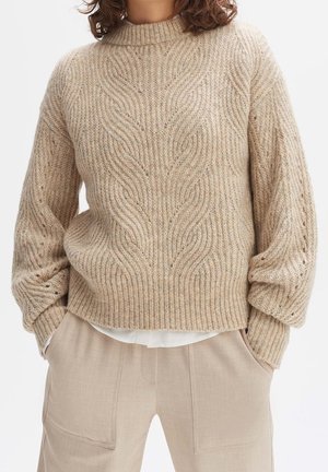 Femme portant un pull à torsades beige et un pantalon beige assorti, les mains dans les poches, debout devant un fond blanc.