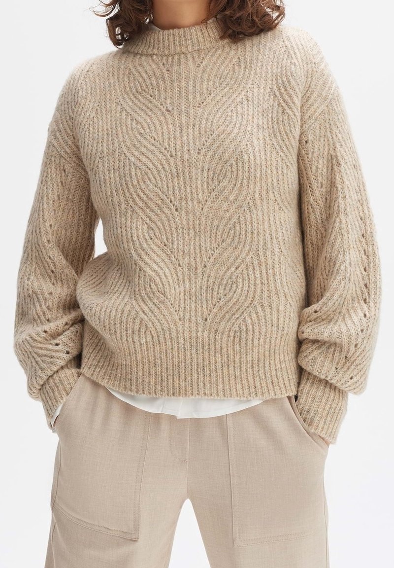 Femme portant un pull à torsades beige et un pantalon beige assorti, les mains dans les poches, debout devant un fond blanc.