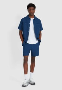 Marineblaues kurzärmeliges Hemd mit Kragen und zwei Taschen, kombiniert mit passenden Shorts. Getragen mit einem weißen T-Shirt und grauen Sneakers.