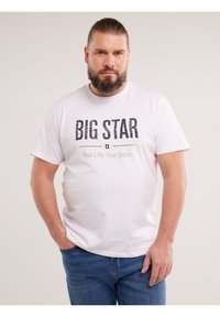 Szakállas férfi hátrafogott hajjal, fehér "BIG STAR" feliratú pólóban és kék farmert viselve, zsebébe dugott kézzel áll egyszerű háttér előtt.