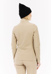 Beige fleecejakke med høj krave, kombineret med en sort ribstrikket hue. Blød tekstur, tætsiddende design og minimale sømme.