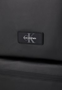Black Calvin Klein bag featuring a smooth texture, rectangular shape, and a branded leather tag with embossed logo. Stitched detailing present.