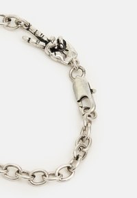 Bracelete de metal prateado com grandes elos ovais e um charme em forma de gesto de mão simbolizando a paz, com fecho de lagosta.