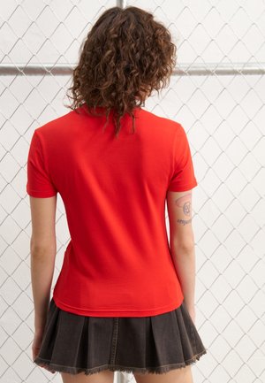 Personne aux cheveux bouclés portant un t-shirt rouge à manches courtes et une jupe plissée noire, debout face à une clôture en grillage.