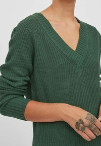Zelený pletený svetr s hlubokým výstřihem do V, texturovanou látkou, dlouhými rukávy a žebrovanými manžetami. Má přiléhavý střih a viditelné tetování.