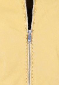 Veste jaune avec une fermeture éclair métallique centrale, texture lisse et design minimaliste, avec une doublure intérieure foncée visible.