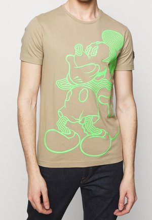 Homme portant un t-shirt beige avec une grande silhouette néon verte d'une souris de dessin animé sur le devant, associé à un jean foncé.