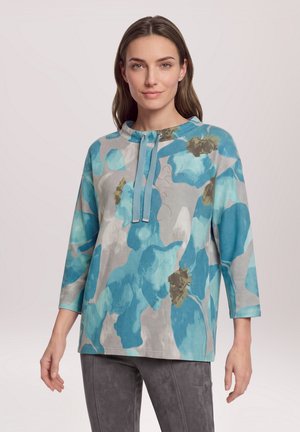 Femme portant un haut à manches longues à motif floral bleu et gris avec col et cordons, associée à un pantalon gris, debout devant un fond uni.