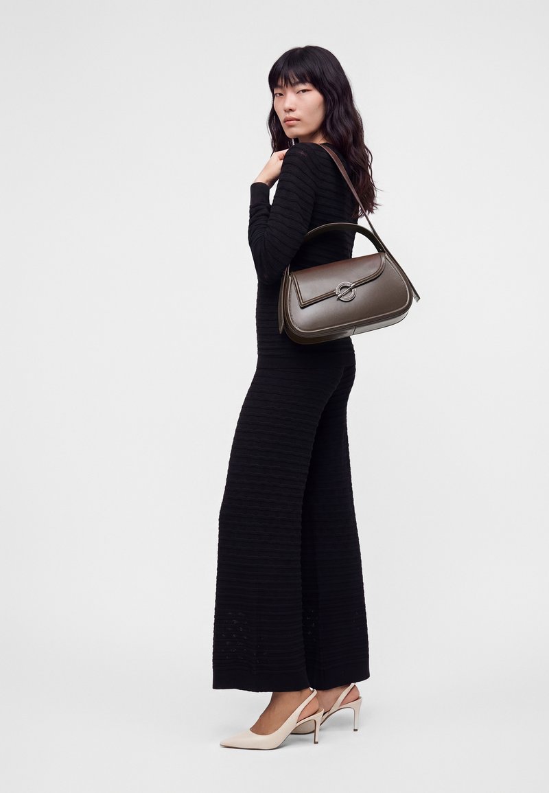 Femme portant une longue robe noire côtelée, des talons beiges et un sac à main en cuir brun foncé avec une fermeture métallique circulaire portée sur l'épaule.