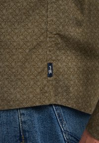 Camisa verde oliva con un patrón geométrico texturizado, que presenta una etiqueta lateral. Combinada con denim, mostrando detalles de tela azul y costuras.
