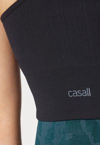 Top de sport noir en tissu côtelé avec un détail de logo argenté, associé à des leggings verts en tricot texturé avec un motif.
