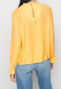 Blusa amarilla de manga larga hecha de tejido texturizado con un escote redondo y detalle de ojal en la espalda. Presenta puños fruncidos y un corte holgado.