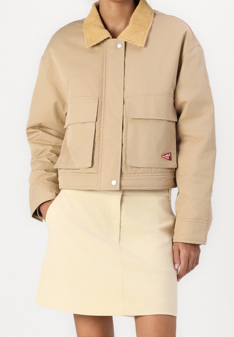 Veste courte beige avec col en velours côtelé et deux grandes poches à rabat devant, portée avec une jupe couleur crème.