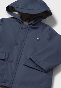 Cappotto impermeabile per bambini di colore blu navy, con cappuccio, bottoni a pressione, polsini regolabili, tasche frontali e un piccolo logo quadrato sul petto.