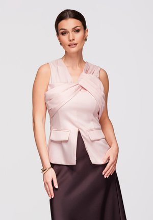 Femme aux cheveux foncés coiffés en un chignon lisse, portant un haut sans manches rose pâle torsadé et une jupe marron foncé, posant avec une main sur la hanche.