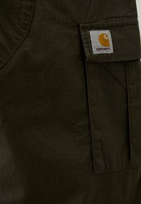 Tissu vert foncé avec un motif de grille subtil, présentant une poche plaquée et une étiquette de logo Carhartt. Le matériau semble durable et texturé.