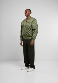 Giacca bomber verde oliva con zip frontale, polsini a costine e tasche. Indossata con pantaloni neri e sneakers bianche su uno sfondo semplice.