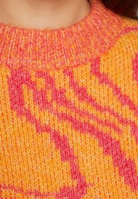 Närbild av en stickad tröjkrage i orange och rosa med en texturerad, ribbad halsringning och abstrakt mönster.