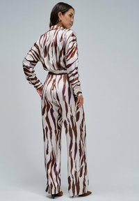 Jumpsuit de seda marrón y crema con patrón abstracto, mangas largas y piernas anchas, con acentos de botones en los puños y cintura ceñida con cinturón.