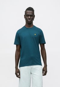 Τιρκουάζ κοντομάνικο μπλουζάκι από βαμβάκι, με κίτρινο κεντημένο λογότυπο στο στήθος και στρογγυλή λαιμόκοψη. Συνδυάζεται με ανοιχτόχρωμο τζιν.