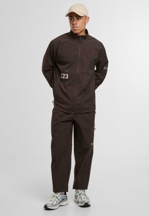 Mand iført en mørkebrun hættejakke med lynlås og bukser med nummer 223, beige kasket og hvide sneakers, stående mod en ensfarvet baggrund.
