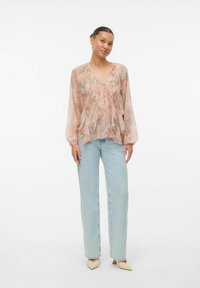 Blusa trasparente con motivo floreale, caratterizzata da ruffles, maniche lunghe e scollatura a V, abbinata a jeans dritti azzurri e tacchi neutri.