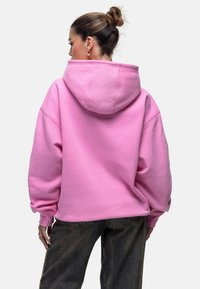 Felpa oversized rosa con cappuccio regolabile, polsini a costine e una morbida texture in pile, abbinata a jeans neri larghi.