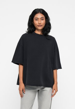 Černé oversized tričko vyrobené z měkké látky, s krátkými rukávy a kulatým výstřihem. Nosené s světlými šedými džíny.
