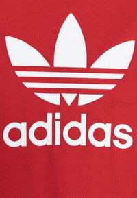 Sarkans kokvilnas T-krekls ar lielu baltu Adidas trīszaru logo un tekstu zem tā treknā, plašā fontā. Dizains ir vienkāršs ar gludām tekstūrām.