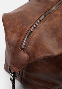 Sac en simili cuir marron avec une fermeture éclair argentée bien visible, de forme arrondie, et des coutures sur la poignée pour un détail supplémentaire.