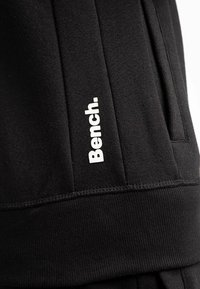 Pantalones de chándal negros hechos de una tela suave, con un logo blanco "Bench." en el costado, con costuras detalladas y bolsillos laterales.