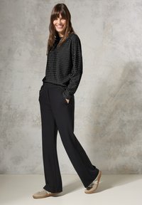 Schwarzer, gemusterter Pullover mit lockerer Passform, kombiniert mit hochgeschnittenen schwarzen Hosen. Das Model trägt beige Sneakers und zeigt einen entspannten Look.