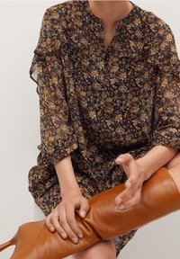 Une personne portant une robe florale à manches longues est assise, une main sur le genou et l'autre ajustant une botte en cuir beige jusqu'au genou.