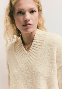 Krémový svetr s texturou a výstřihem do V s proužkovými detaily, vyrobený z měkkého materiálu. Doplňky zahrnují zlatý řetízek s přívěskem.