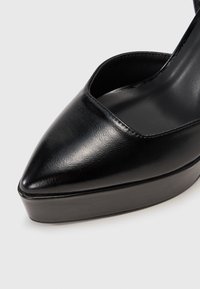 Musta nahkakengät, joissa on korkea korko ja terävä kärki, sileä pinta ja paksu alusta lisäämään korkeutta. Minimalistinen muotoilu, jossa on leikattuja yksityiskohtia.