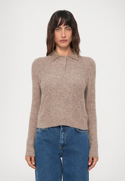 Femme portant un pull en tricot beige avec un col et des boutons, associé à un jean bleu taille haute, se tenant contre un fond uni.