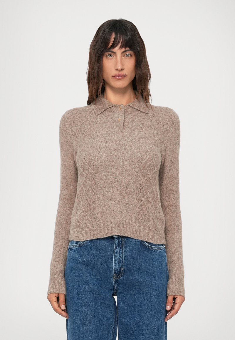Mujer vistiendo un suéter de punto beige con cuello y botones, combinado con jeans de tiro alto azules, de pie frente a un fondo plano.