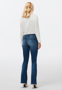 Jean flare en denim bleu avec une taille mi-haute, présentant un design classique, cinq poches et une texture lisse. Blouse blanche à manches longues.