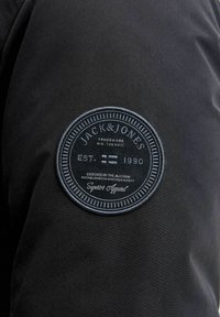Sort stofjakke med en rund broderet patch med "JACK & JONES," etableringsår "EST. 1990," og dekorative detaljer.