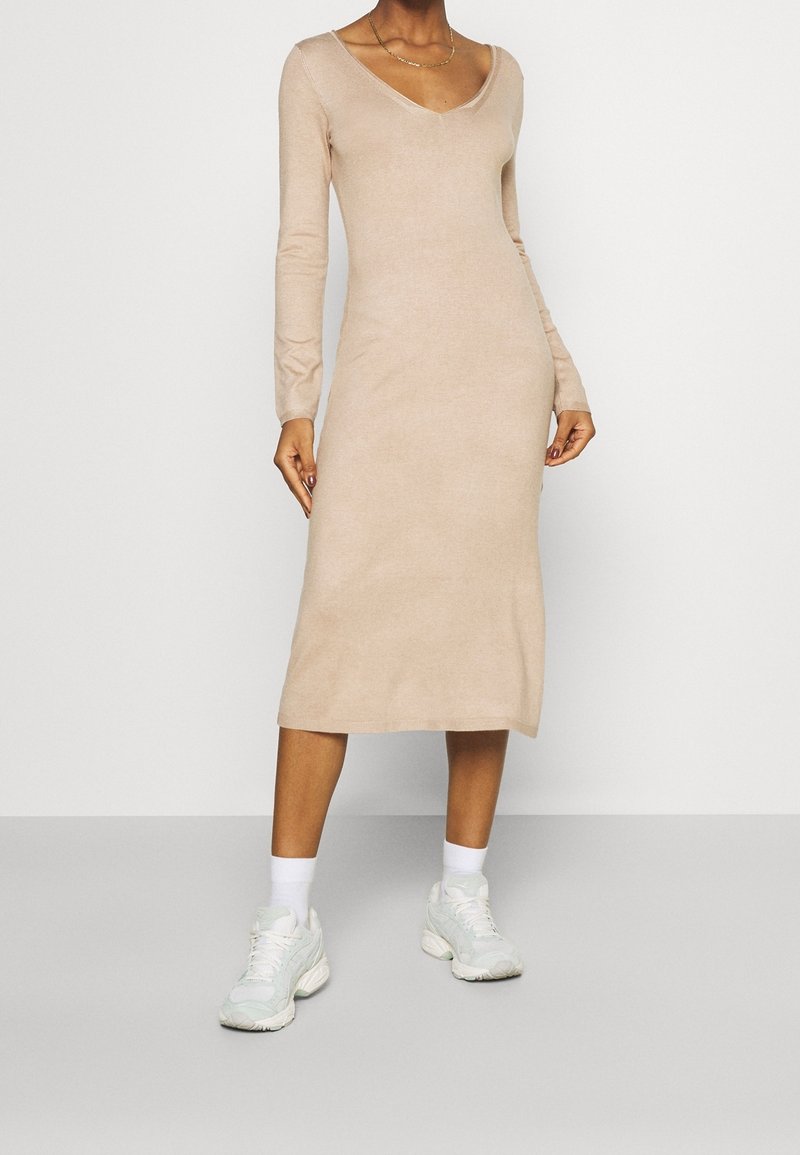 Femme portant une robe midi beige à manches longues associée à des chaussettes blanches et des baskets blanches épaisses, debout devant un fond uni.