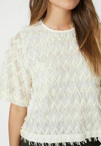 White sheer top featuring a zigzag pattern with fringed details, short sleeves, and a round neckline. Textured fabric adds visual interest.