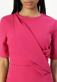 Robe fuchsia avec un col rond et des manches courtes. Présente un détail drapé à l'avant et une texture lisse. Aucun motif ni hardware visible.