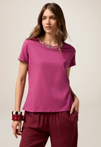 T-shirt fuchsia à manches courtes, avec un col fleuri d�écoratif et coloré. Associé à un pantalon bordeaux fluide et à des bracelets empilés.