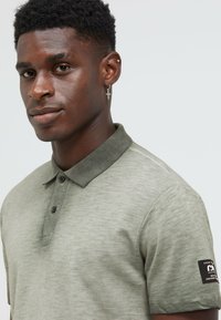 Kurzärmliges olivgrünes Polohemd mit strukturiertem Finish, einem grauen Kragen und einem schwarzen Etikett am linken Ärmel. Drei-Knopf-Leiste.