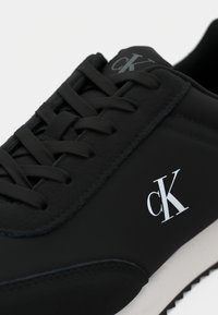 Black sneaker with a matte finish, featuring flat laces, a white logo, and a textured sole. Clean design with subtle accents.