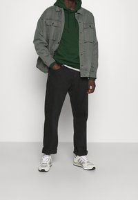 Grijze spijkerjasje over een groene hoodie, zwarte jeans en grijze sneakers met witte en olijfgroene accenten. Casual gelaagde outfit.