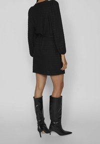 Una persona está de pie en un vestido negro texturizado ajustado en la cintura, combinado con tacones negros hasta la rodilla, frente a un fondo neutro.