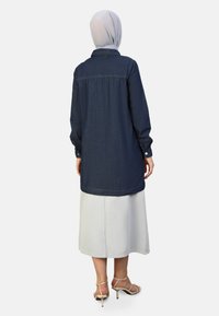 Donkere blauwe spijkerjas met een knoopsluiting aan de voorkant, lange mouwen en een gebogen zoom, gecombineerd met een lichtgrijze rok. Vlotte texturen en een getailleerde pasvorm.