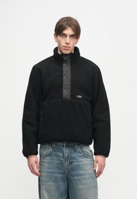 Μαύρο φλις πόλο με ψηλό κολάρο και κλείσιμο με κουμπιά snap μπροστά. Περιλαμβάνει πλαϊνές φερμουάρ και υφή στην επιφ�άνεια.