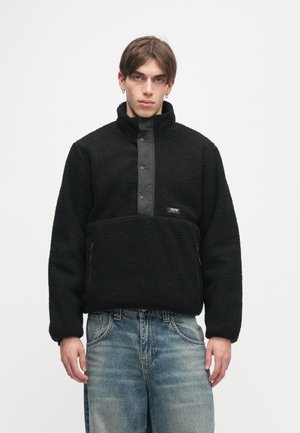 Polaire preto com gola alta e fecho de botão de pressão na frente. Apresenta zíperes laterais e uma superfície texturizada.